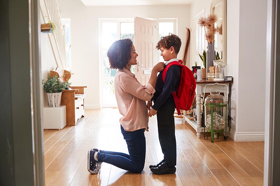 Personal Insurance - Mom Kneels in a Large Hallway to Straighten Her Son's Red Backpack on His Shoulders, Front Door Open and Sun Shining Through